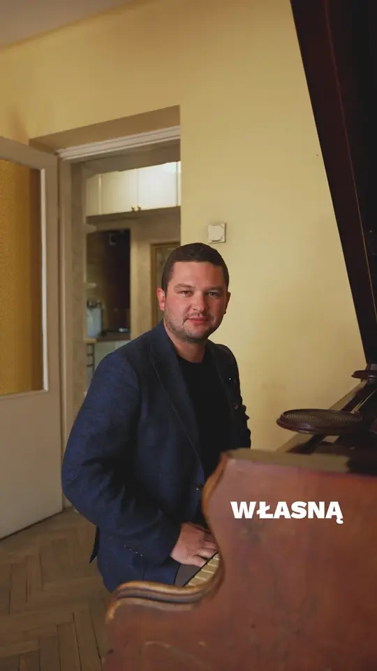 Artur Zając w granatowej marynarce przy fortepianie w pokoju z żółtymi ścianami i parkietem w jodełkę