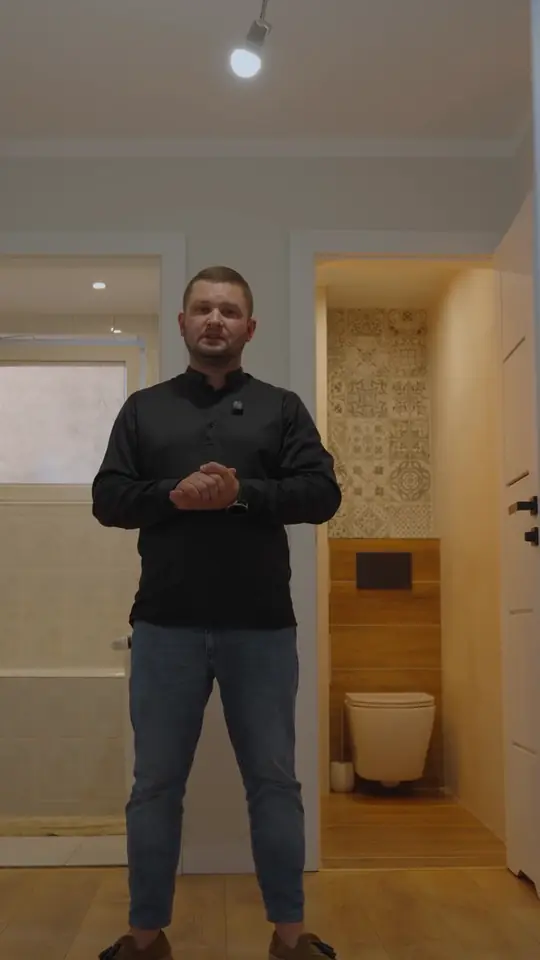 Mężczyzna w czarnej koszuli i dżinsach w jasnym przedpokoju, za nim łazienka z podwieszaną toaletą