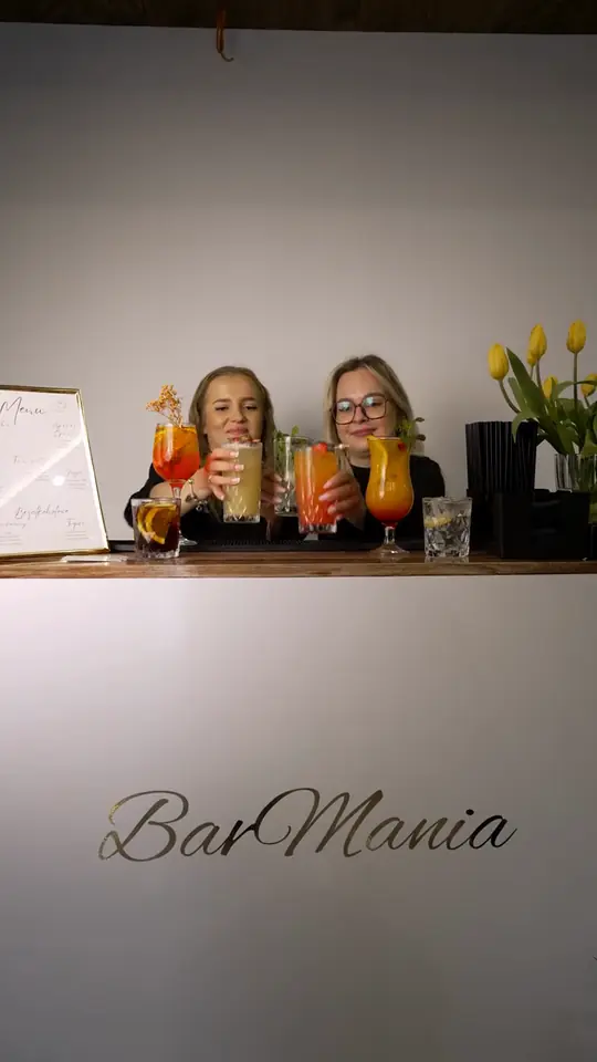 Dwie kobiety za białym barem z logo BarMania trzymają drinki, obok menu w ramce i żółte tulipany