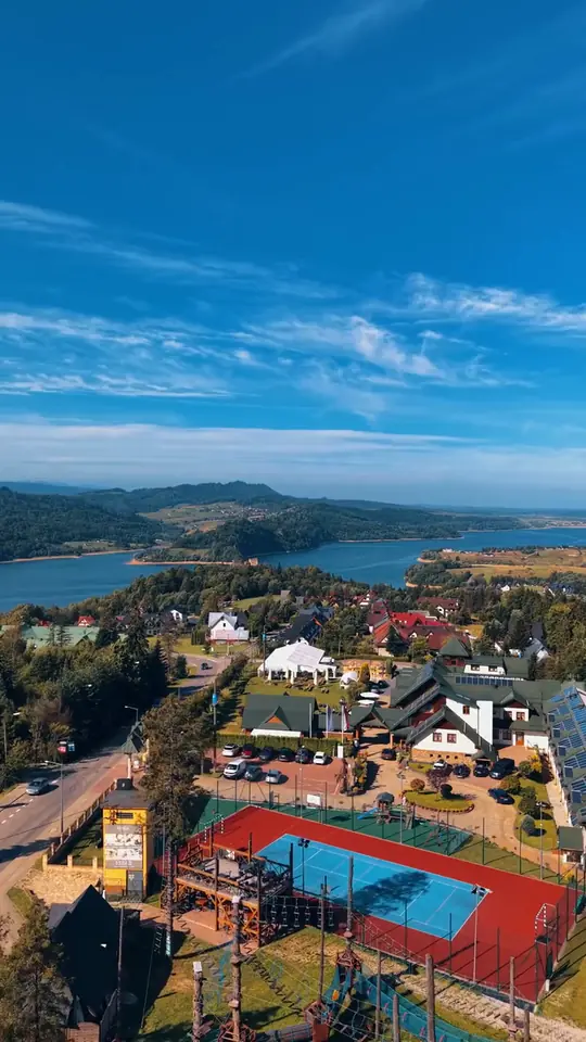 Widok z drona na resort z kortem i zielonymi dachami, za nim jezioro i zalesione wzgórza