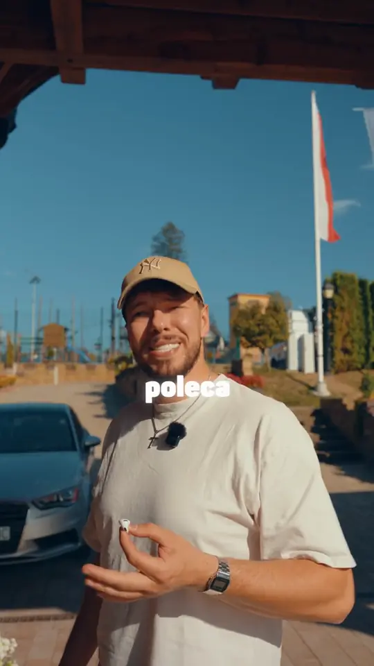 Uśmiechnięty mężczyzna w beżowej czapce trzyma słuchawkę douszną, za nim podjazd, flaga i zaparkowane auto