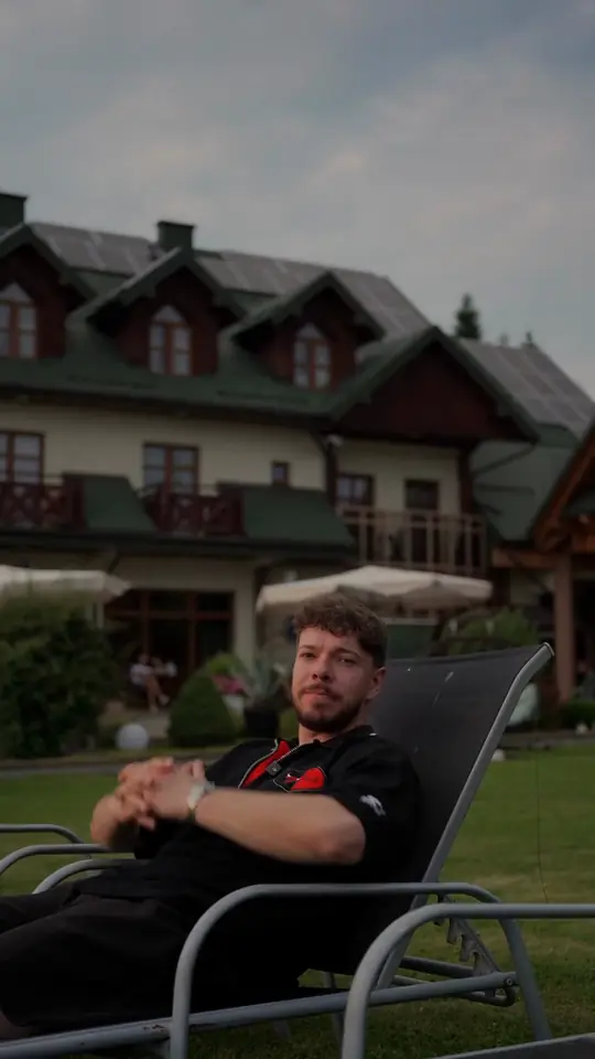 Mężczyzna w czarnej koszulce polo na leżaku przed budynkiem resortu z drewnianymi balkonami