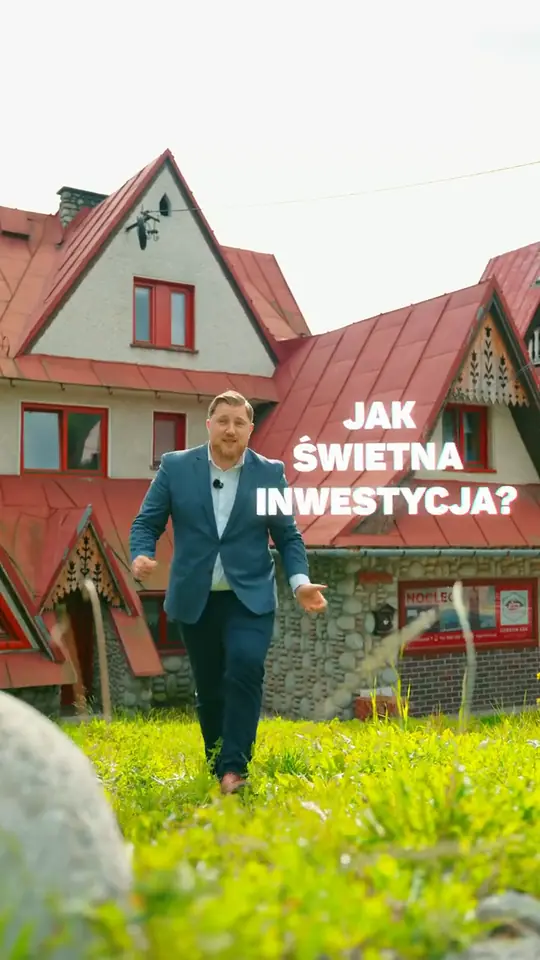 Jan Gryguś w niebieskiej marynarce idzie po trawie przed budynkiem z czerwonym dachem i kamienną podmurówką