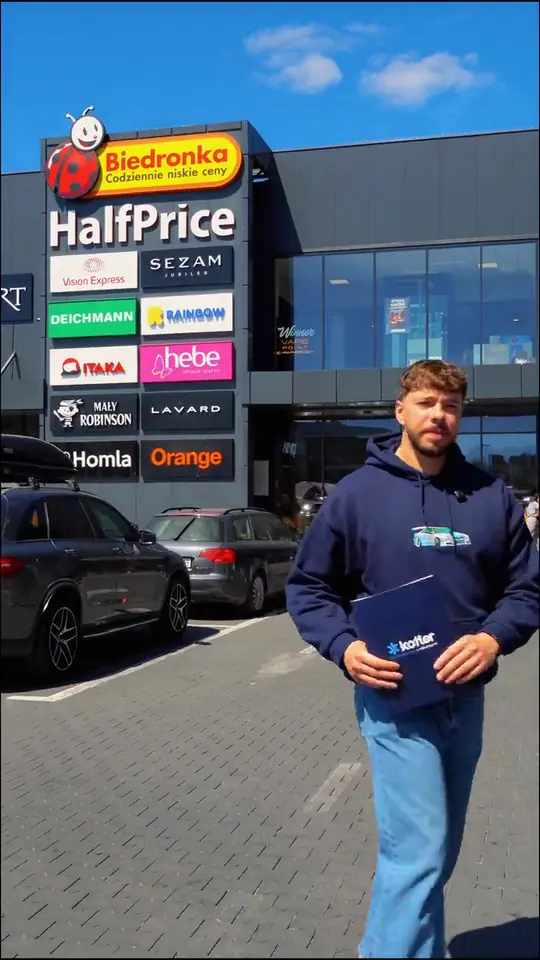 Mężczyzna z teczką z logo Kotler idzie przez parking przed Stopiak Parkiem Handlowym, w tle szyldy sklepów