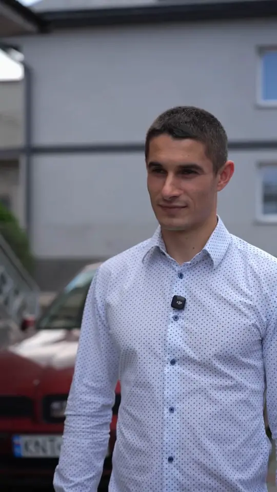 Agent w białej koszuli w kropki stoi na ulicy przed szarym domem, za nim czerwony samochód