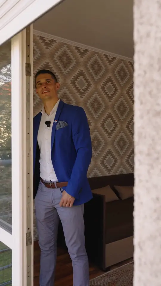Agent w niebieskiej marynarce stoi w drzwiach balkonowych pokoju ze wzorzystą tapetą i brązową sofą