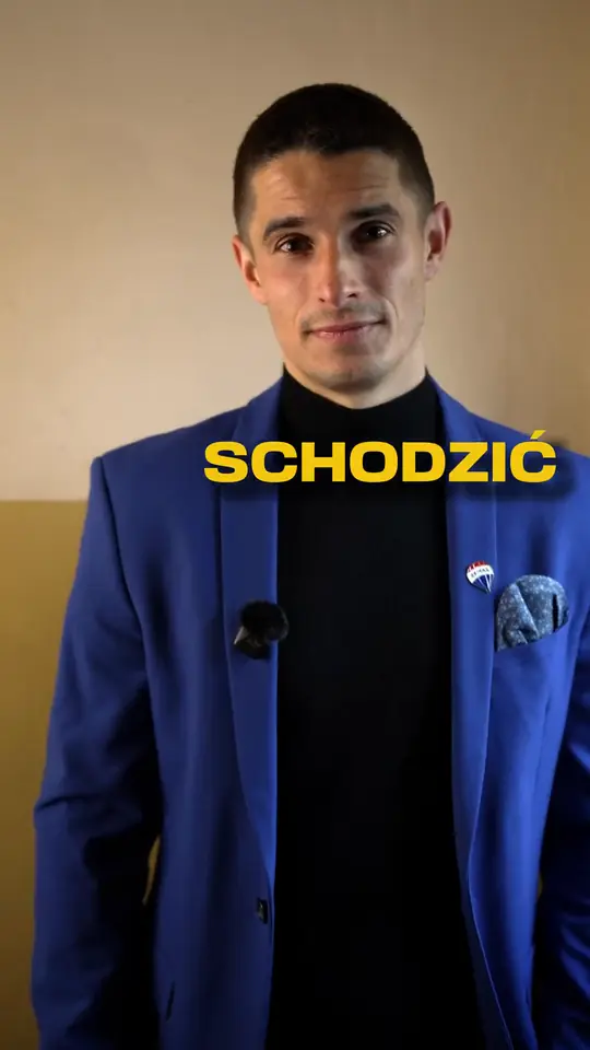 Agent w niebieskiej marynarce i czarnym golfie z przypinką RE/MAX patrzy w kamerę, obok żółty napis SCHODZIĆ