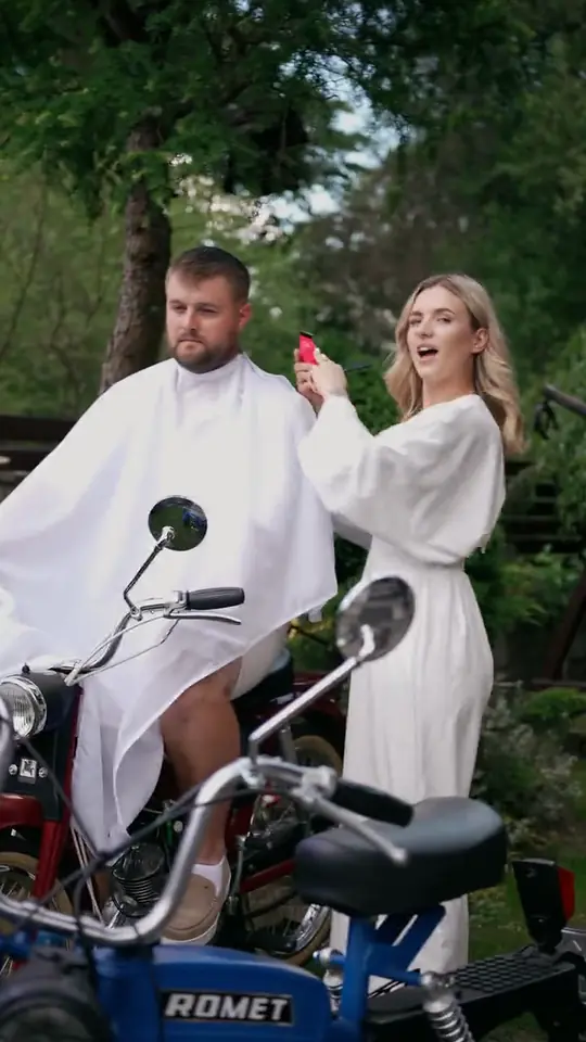 Barberka w bieli strzyże maszynką mężczyznę w pelerynie siedzącego na motorowerze w ogrodzie