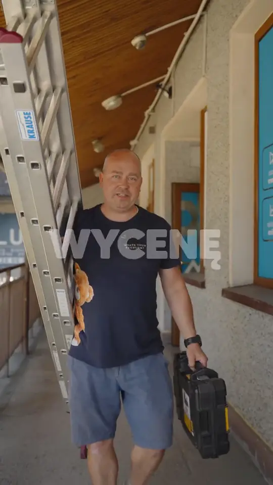 Mężczyzna z aluminiową drabiną na ramieniu i czarną skrzynką narzędziową idzie zadaszonym korytarzem