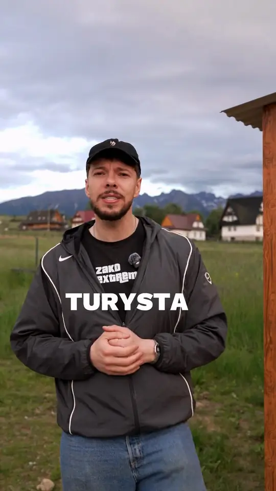 Mężczyzna w czapce, kurtce i koszulce z logo ZakoExtreme mówi na łące, za nim domy i góry, na kadrze napis TURYSTA