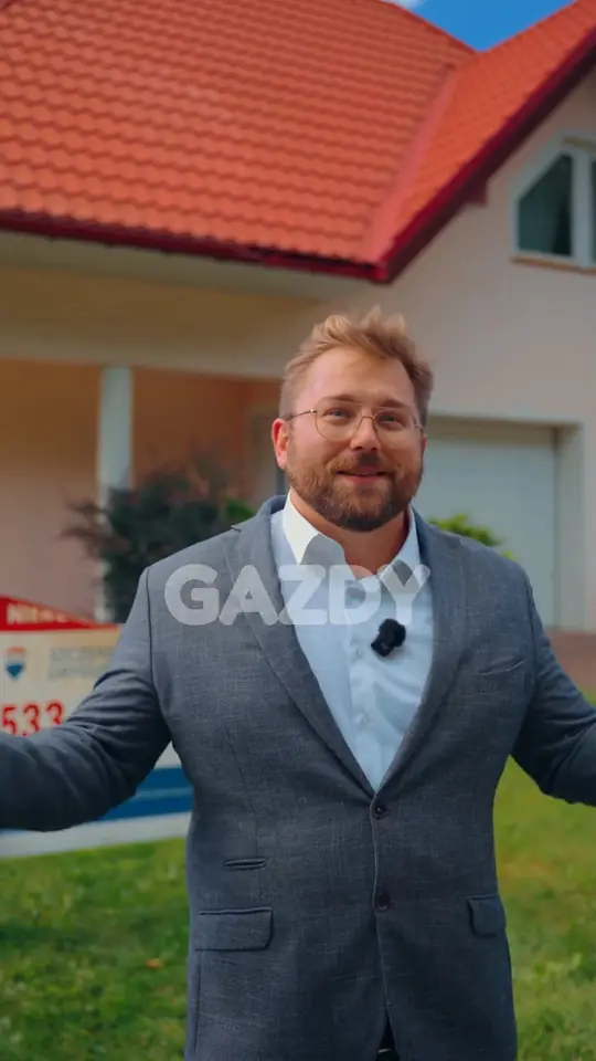 Szczepan Gryguś w szarym garniturze z rozpostartymi rękami przed domem z czerwonym dachem, obok baner RE/MAX
