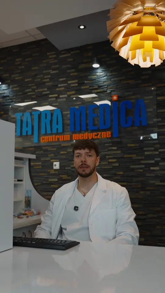Mężczyzna w białym fartuchu za ladą recepcji, za nim logo Tatra Medica centrum medyczne na kamiennej ścianie