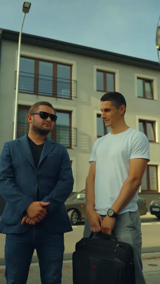 Dwóch mężczyzn przed jasnym blokiem, jeden w marynarce i okularach przeciwsłonecznych, drugi w białym T-shircie z torbą