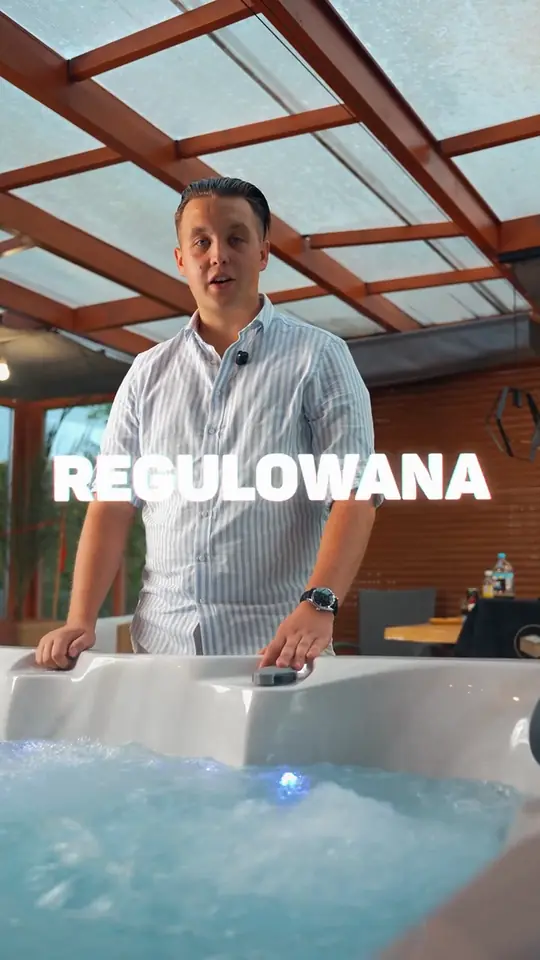 Mężczyzna w koszuli w paski przy białym jacuzzi z bulgoczącą wodą, pod przeszklonym dachem na drewnianych belkach