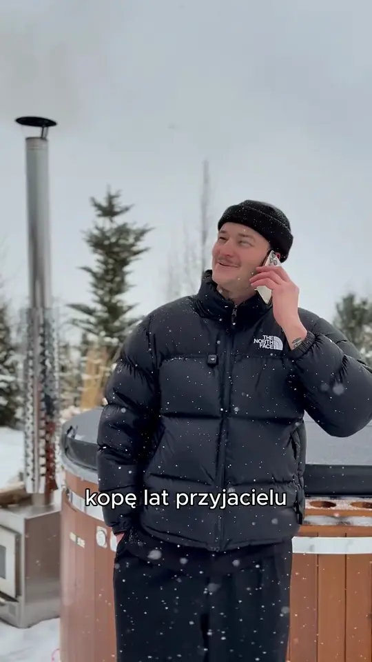 Mężczyzna w czarnej kurtce i czapce rozmawia przez telefon przy drewnianej balii z kominem, pada śnieg